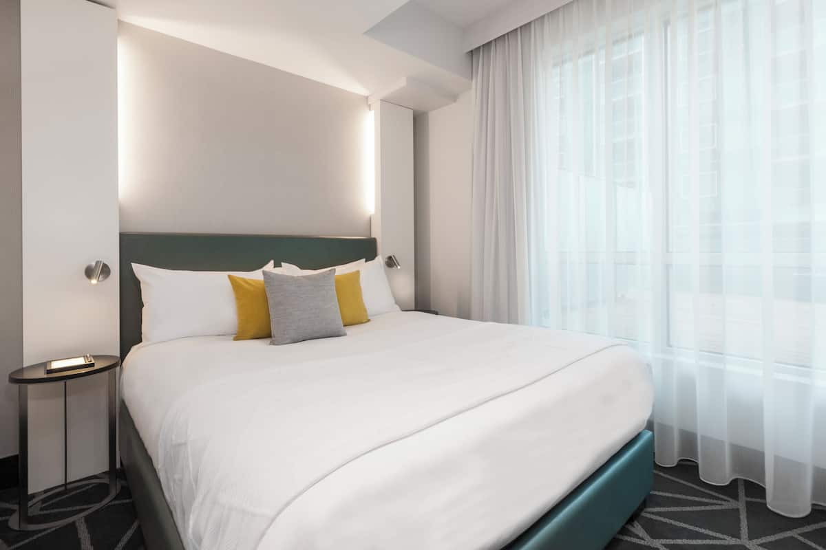 Una habitación de hotel moderna con una cama grande, una mesita de noche y una ventana con cortinas traslúcidas.