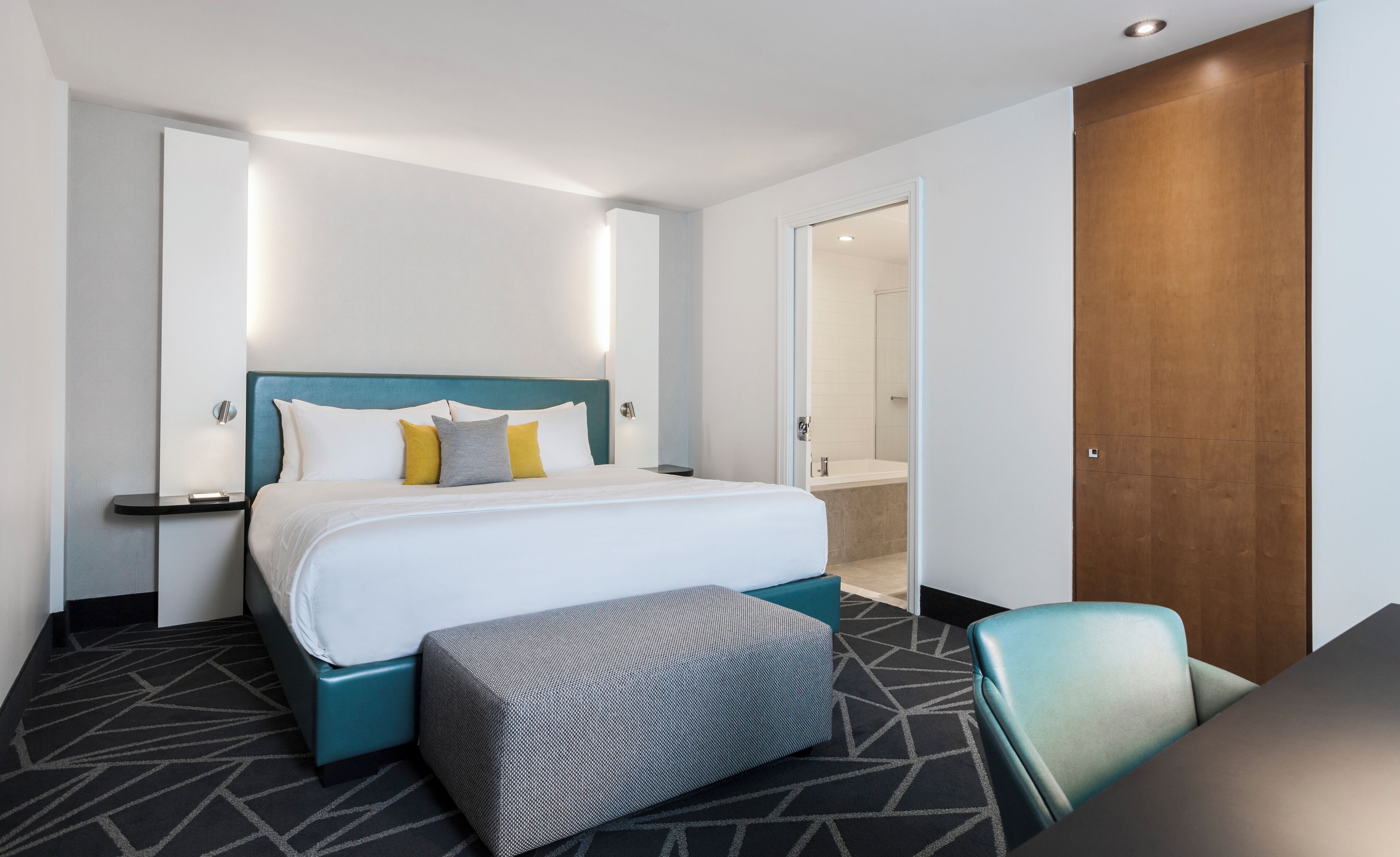 Habitación de hotel con una cama grande, una mesita de noche, una silla y un baño visible a través de una puerta abierta.