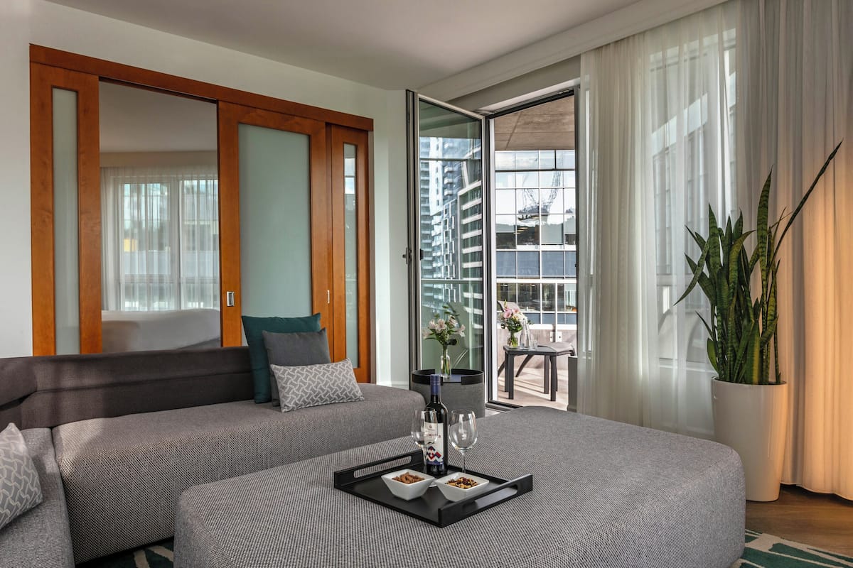 Una habitación de hotel moderna con sofá, mesa de comedor con bandeja de comida y bebidas, ventana grande con cortinas y una planta en maceta.