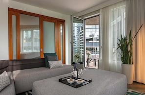Una habitación de hotel moderna con sofá, mesa de comedor con bandeja de comida y bebidas, ventana grande con cortinas y una planta en maceta.