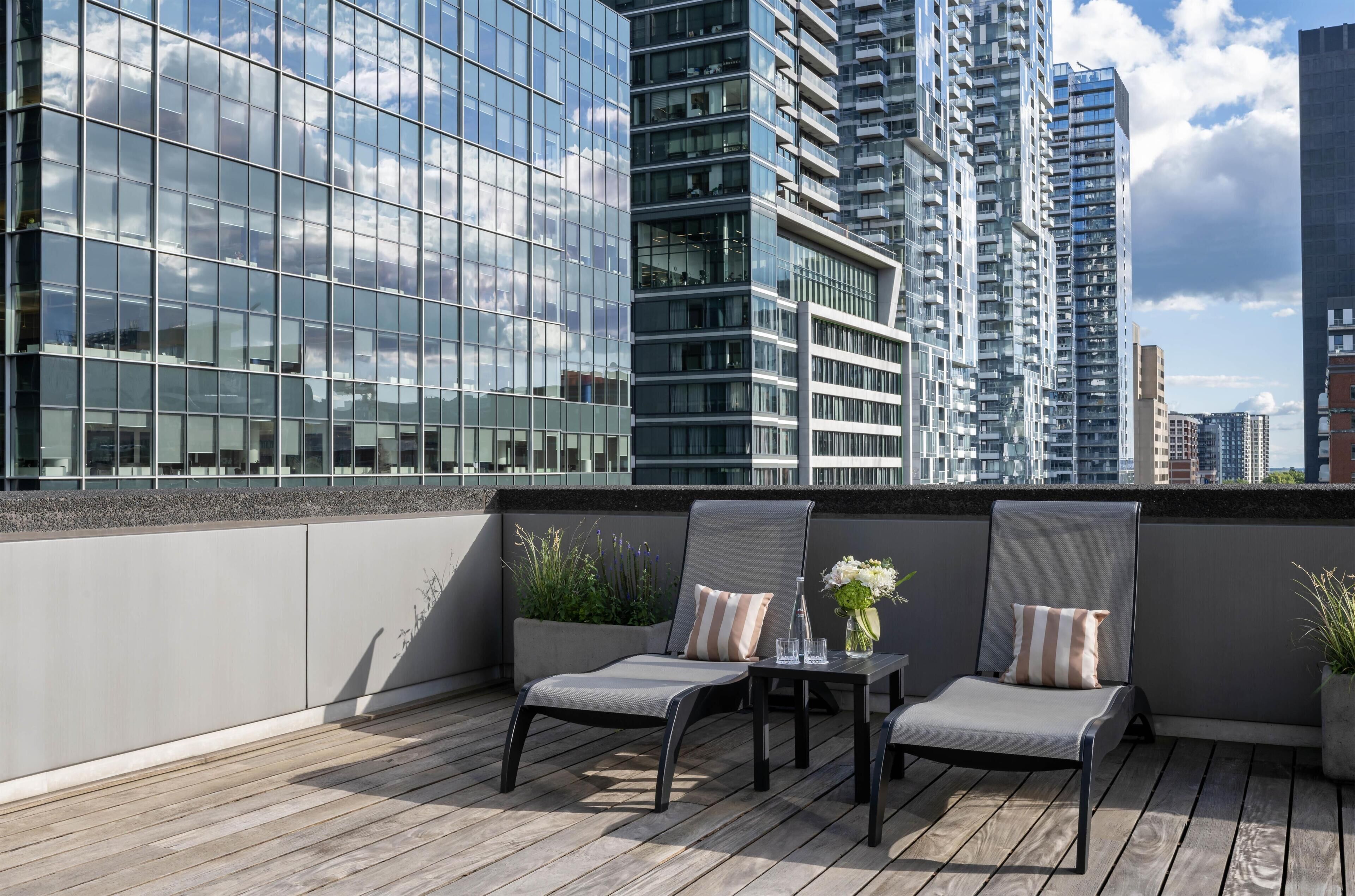 A rooftop terrace with two chairs and a table, overlooking a cityscape with modern buildings.