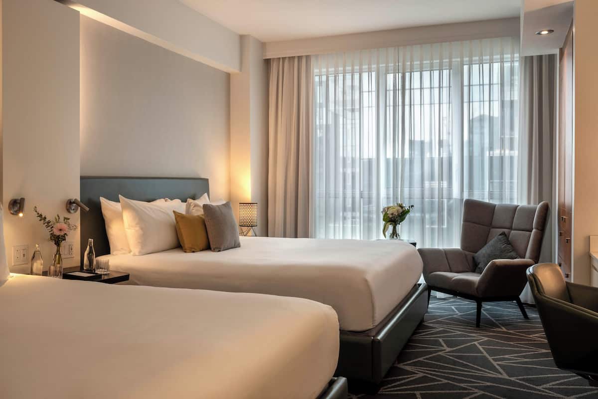 Habitación de hotel con dos camas, una silla, un escritorio y un amplio ventanal con cortinas.