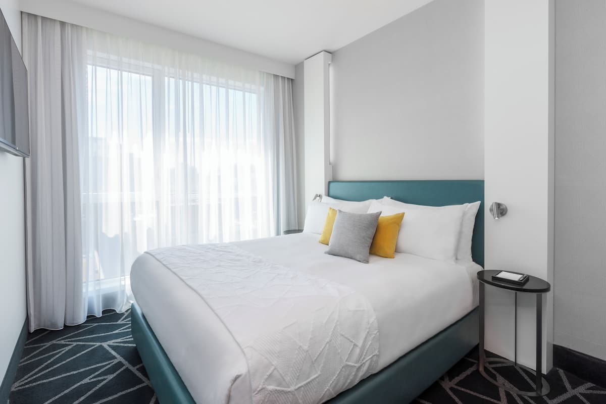Una habitación de hotel con una cama grande, una mesita de noche, un televisor en la pared y una ventana con cortinas translúcidas.