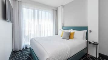 A hotel room with a large bed, a bedside table, a TV mounted on the wall, and a window with sheer curtains.