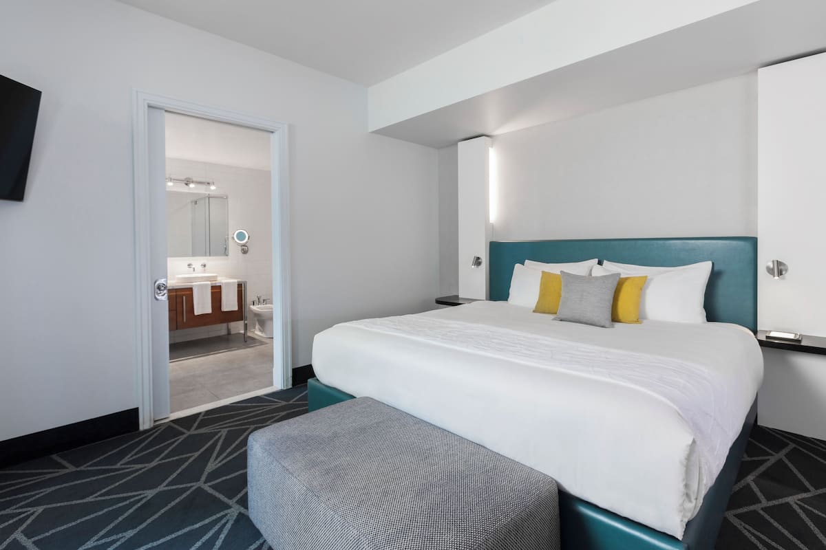 Habitación de hotel con cama, banca, mesita de noche, televisor y baño visible a través de una puerta abierta.