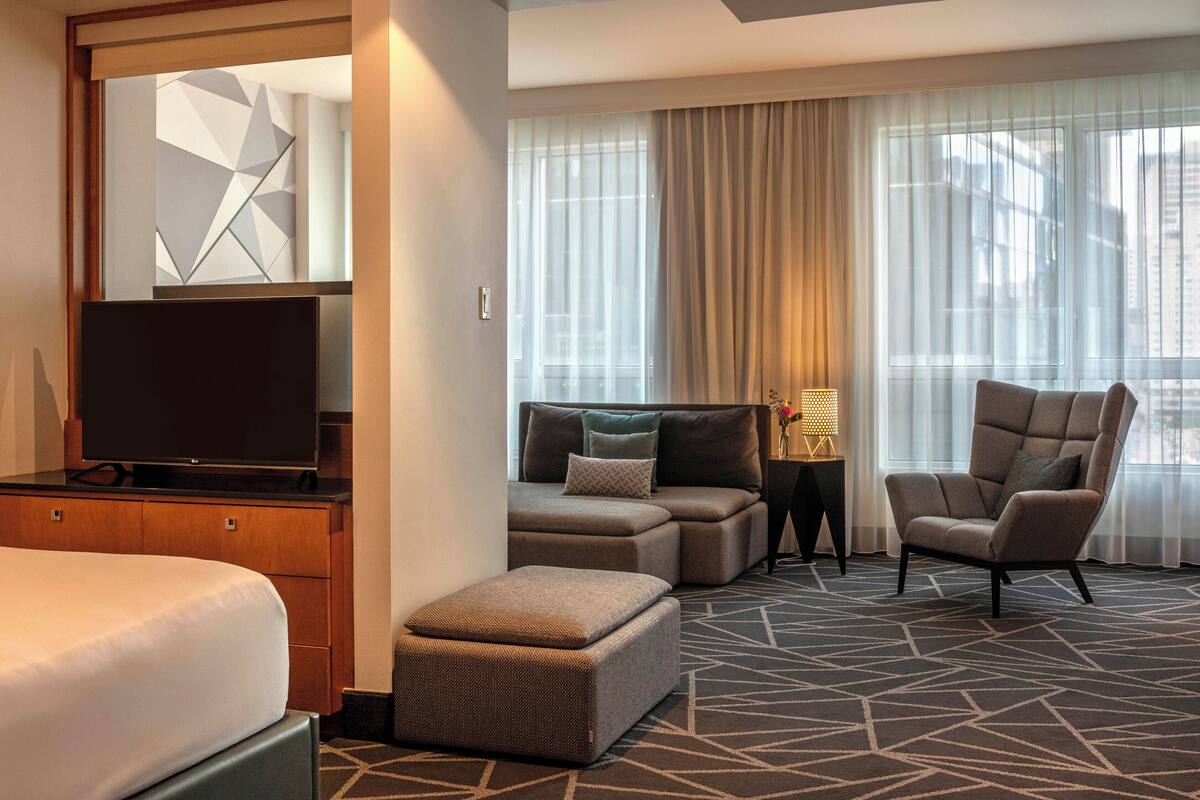 Una habitación de hotel con cama, televisión, sofá, sillón y una mesita con lámpara.