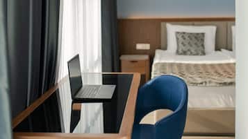 A modern hotel room with a wooden desk, a laptop, a blue chair, a bed with white bedding, and a hanging light fixture.