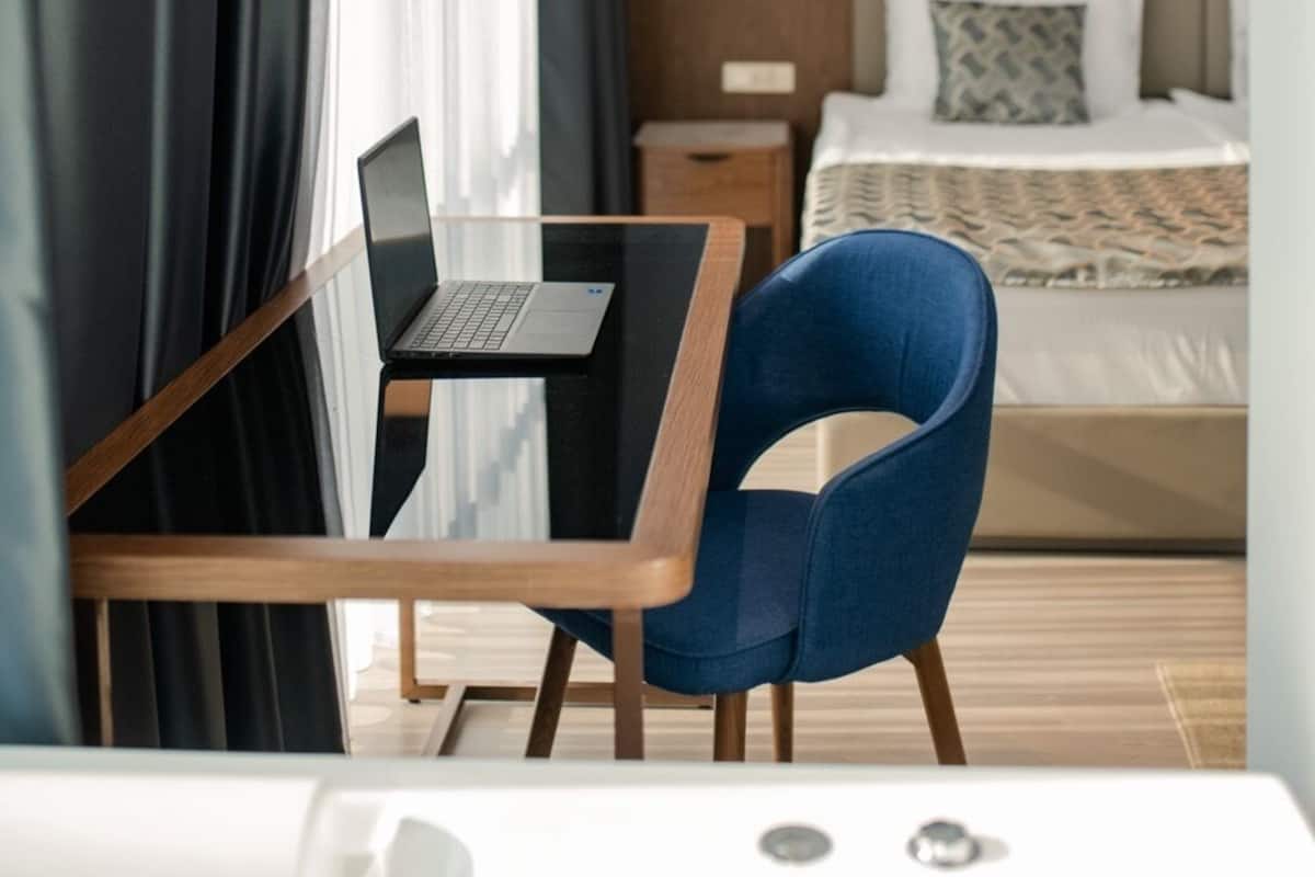 A modern hotel room with a wooden desk, a laptop, a blue chair, a bed with white bedding, and a hanging light fixture.