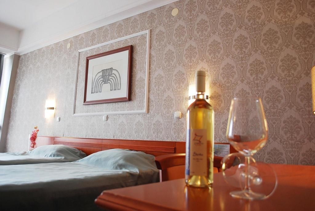 Camera d'albergo con un letto, un tavolo con bottiglia e bicchiere, un quadro appeso al muro e carta da parati decorata.