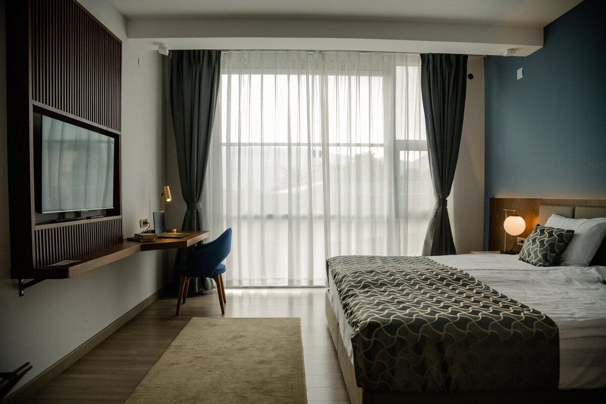 A hotel room with a bed, a desk with a chair, a TV, and a window with curtains.