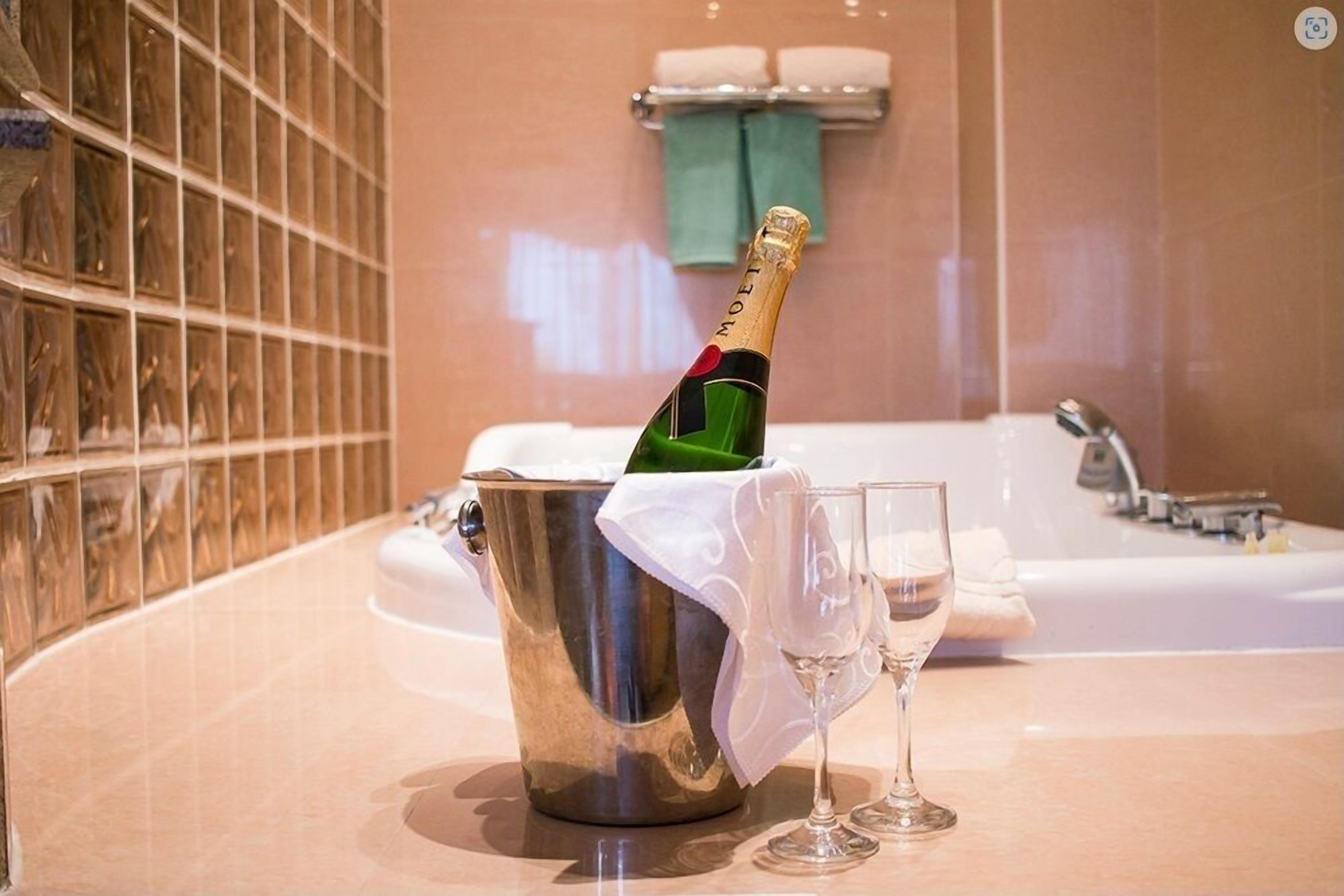 A champagne bottle in a metal bucket with two glasses, a bathtub, and a tiled wall in a bathroom.