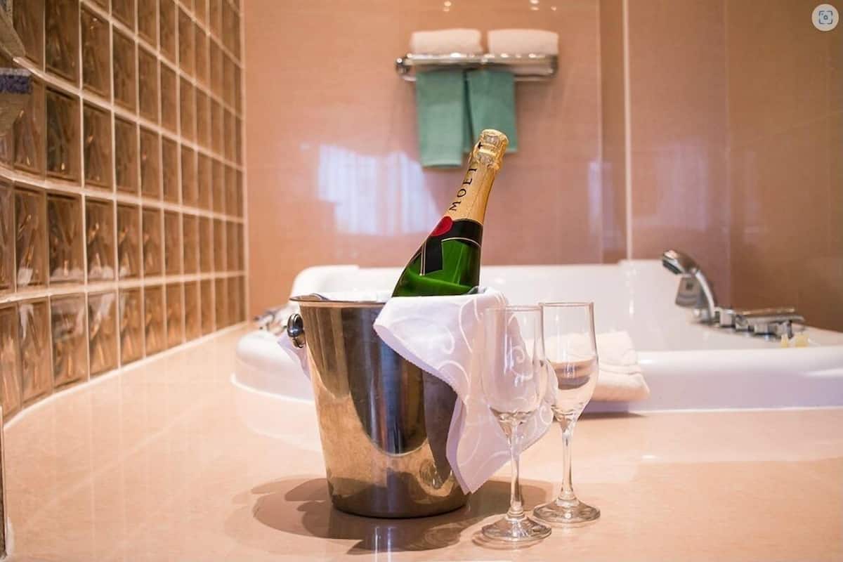 A champagne bottle in a metal bucket with two glasses, a bathtub, and a tiled wall in a bathroom.