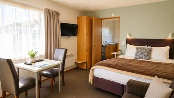 A hotel room with a bed, a desk with a chair, a TV, a wardrobe, and a small table with a plant.