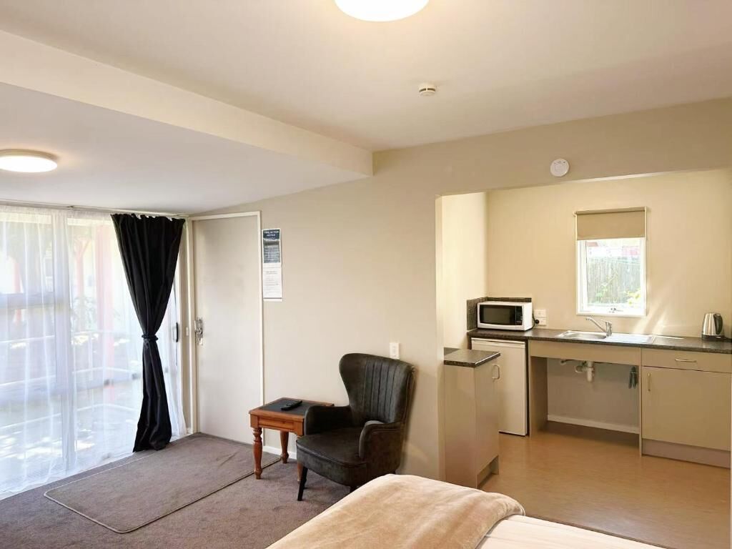 A compact room with a bed, a chair, a small table, a kitchenette with a microwave, and a window.