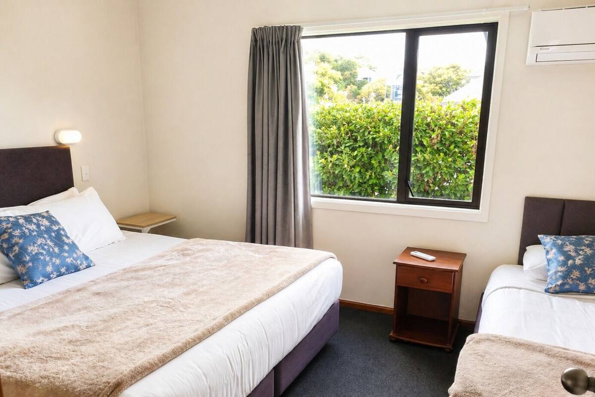 A hotel room with two beds, a window with a view of greenery, a bedside table with a remote control, and a wall-mounted air conditioning unit.