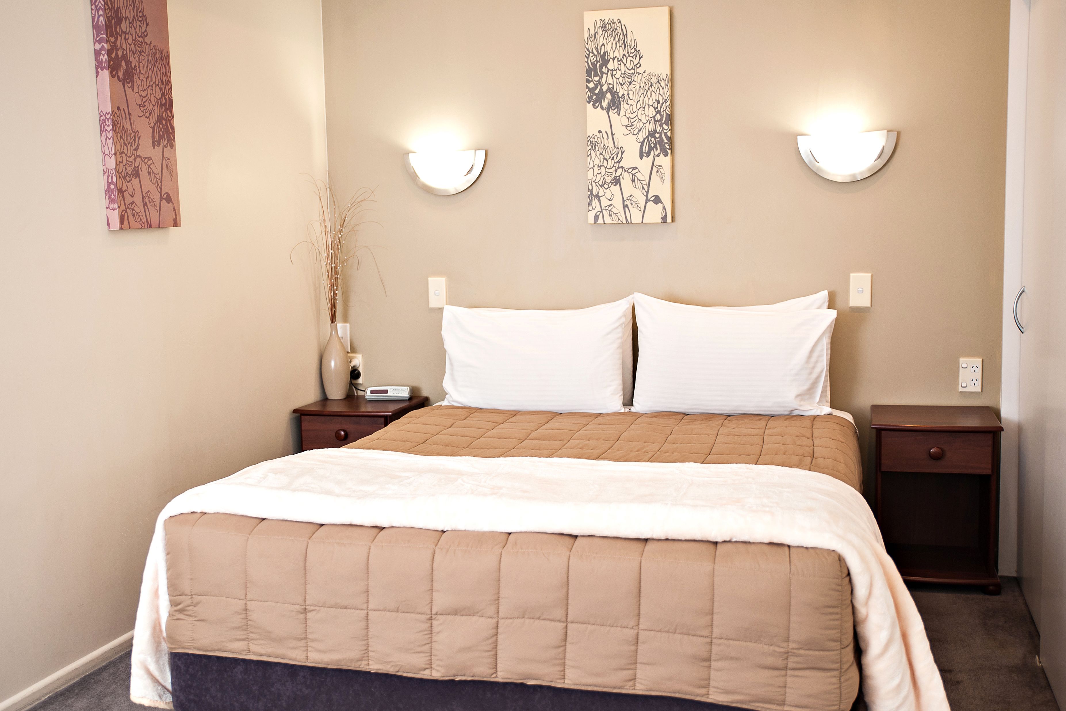 Una cama bien hecha con un edredón color beige, dos mesitas de noche con lámparas, y una obra de arte en la pared sobre la cama.