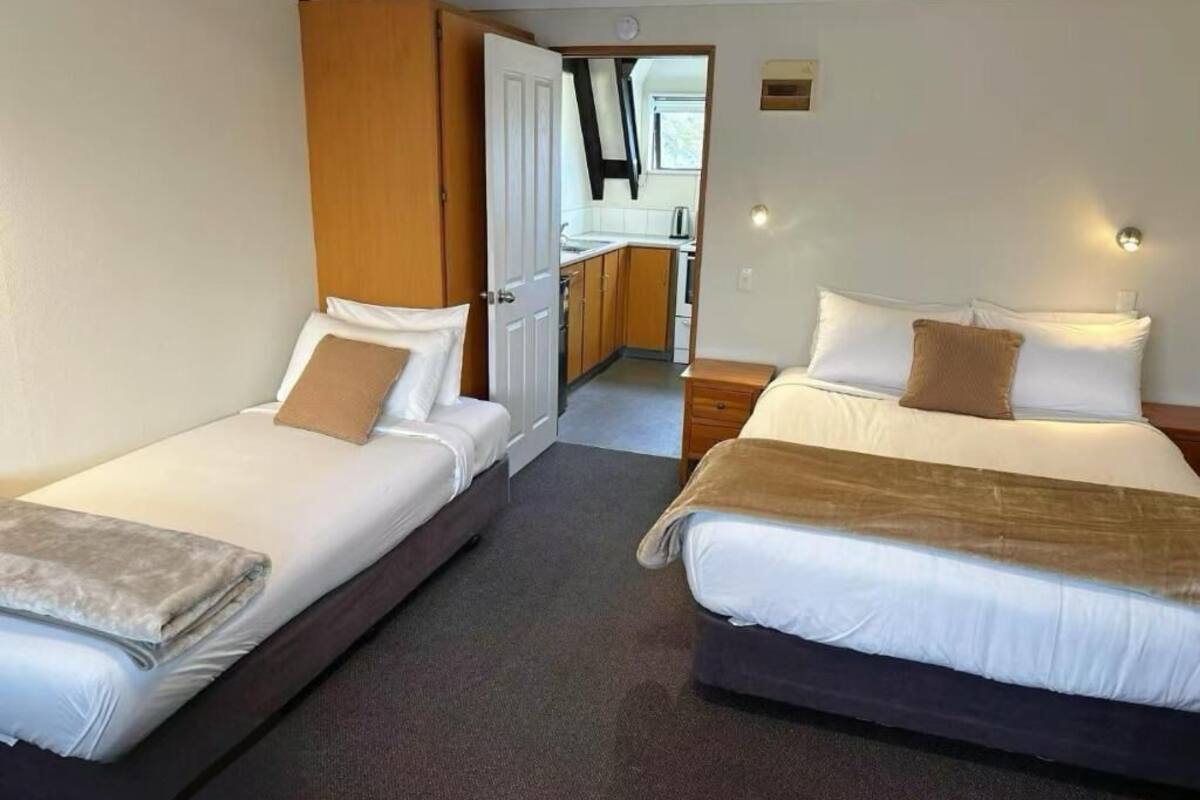 Two single beds with pillows, a wooden wardrobe, a small kitchen area, and a door leading to a balcony.
