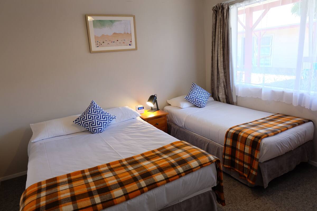 Two single beds with white linens and plaid blankets, a nightstand with a lamp, a framed picture on the wall, and a window with curtains.