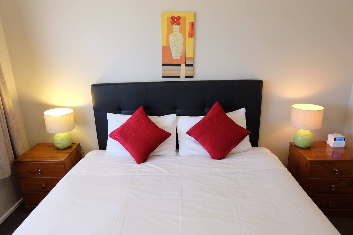 A neatly made bed with white bedding and two red pillows, flanked by wooden nightstands with lamps.
