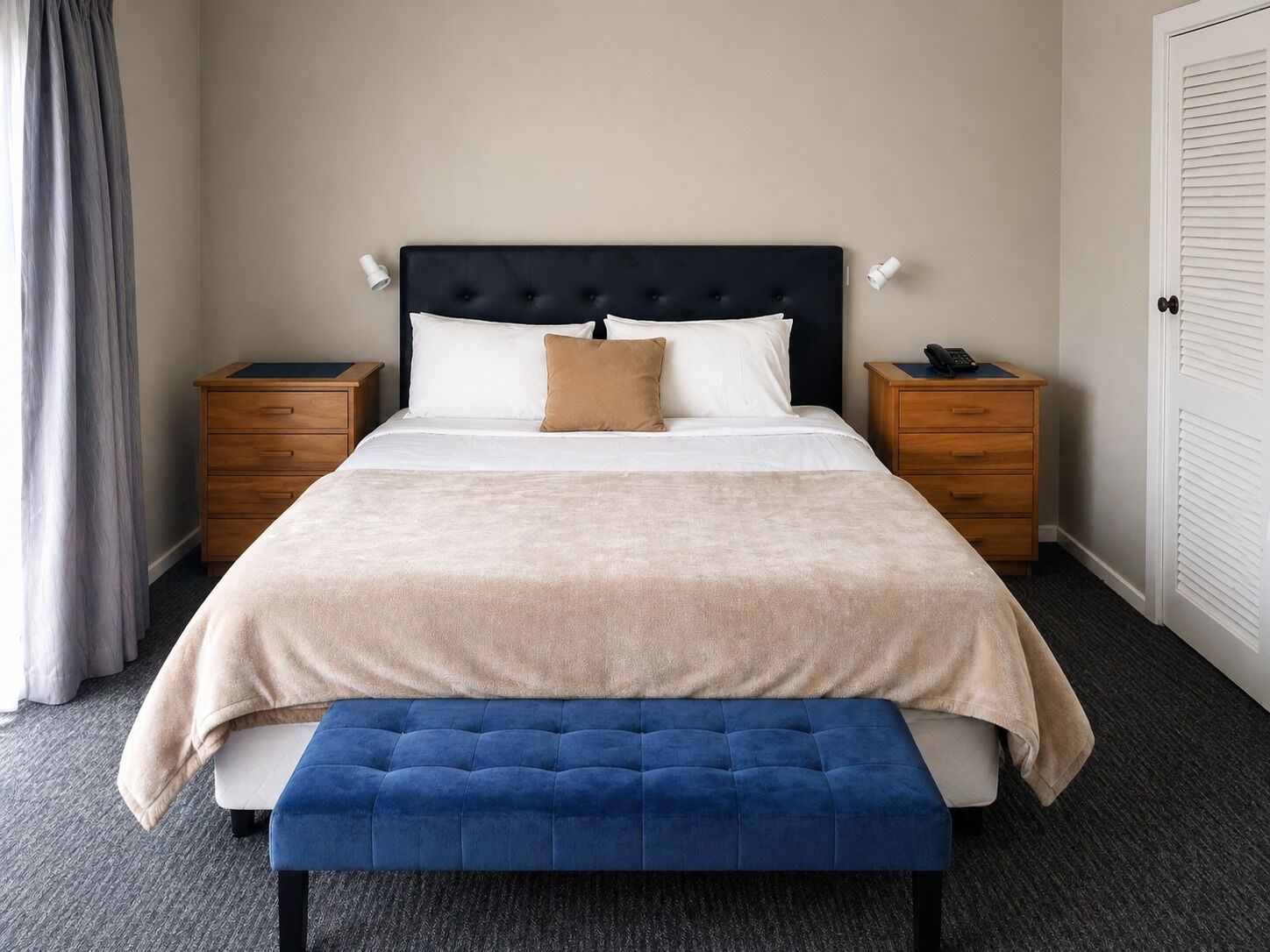 A neatly made bed with a headboard, two bedside tables, a bench, and a door.