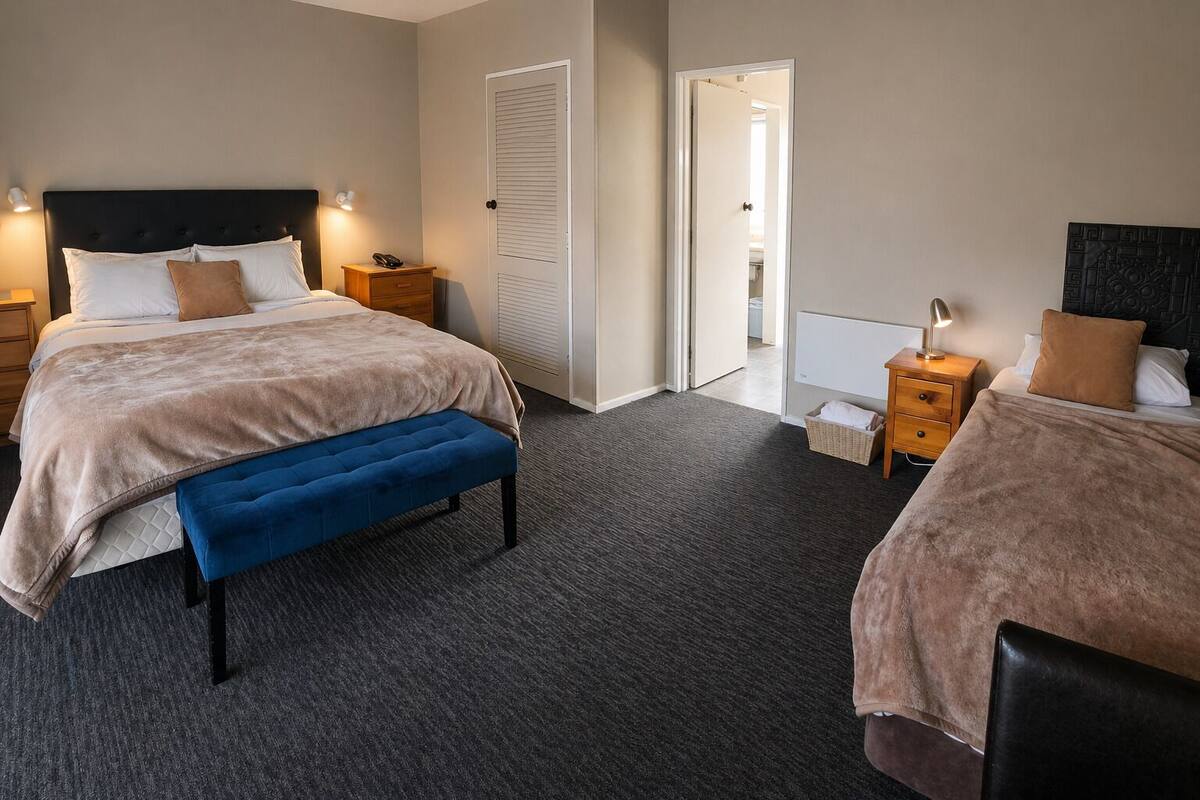 A hotel room with a large bed, a smaller bed, a bench, two bedside tables, and a door leading to another room.