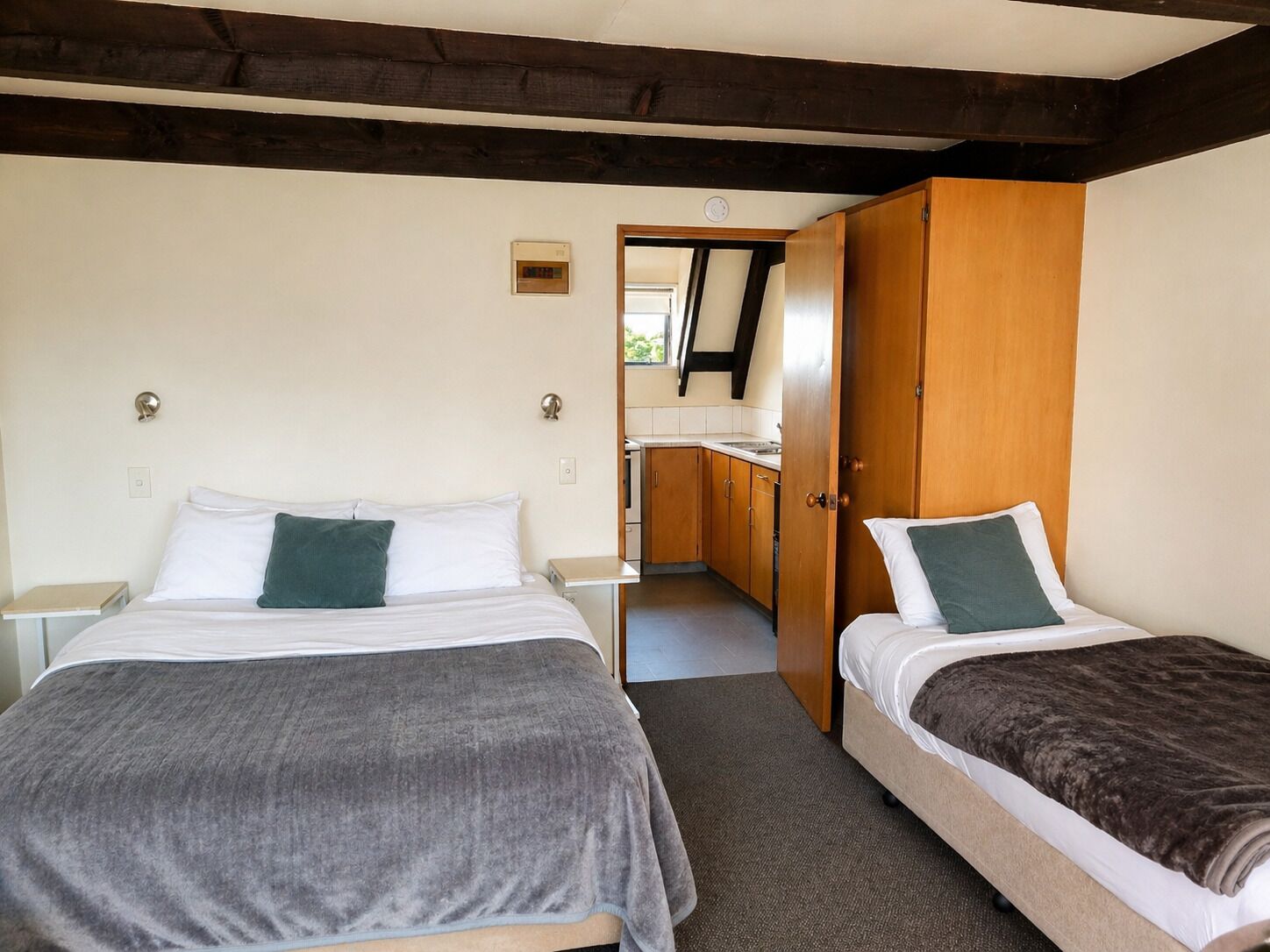 A room with two beds, a wooden wardrobe, a small kitchenette, and a door leading to another room.