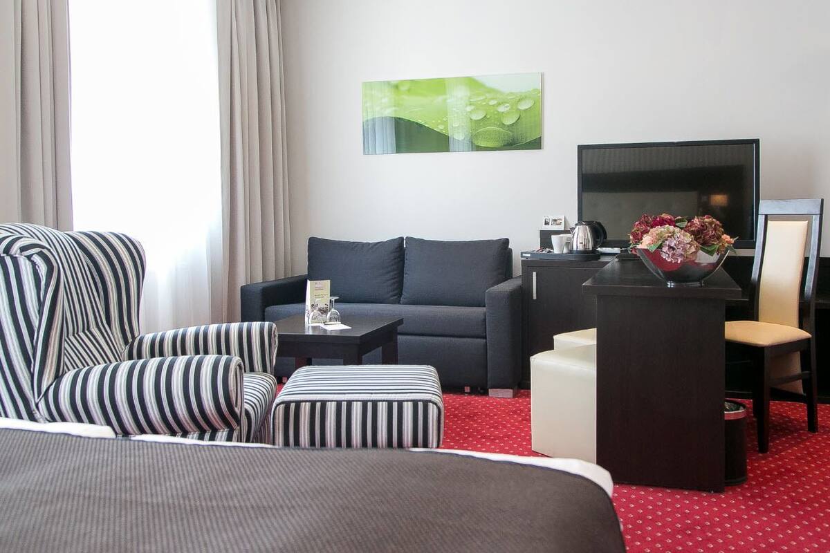 A hotel room with a bed, a striped armchair, a sofa, a small table with a vase of flowers, a desk with a chair, and a TV on the wall.