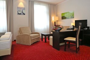 Una habitación de hotel con alfombra roja, un sillón color beige, una pequeña mesa con un florero y un escritorio con una computadora y un televisor.