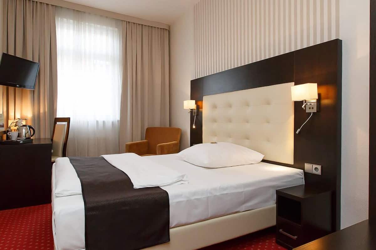 A hotel room with a large bed, a TV, a chair, and a desk with a lamp.