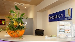A reception desk with a sign reading "Rezeption" and a Hotel Herbst logo. There is a large potted plant on the desk, a black chair, and a bulletin board with various papers and a brochure. - Hotel Herbst Berlin (Berlin)