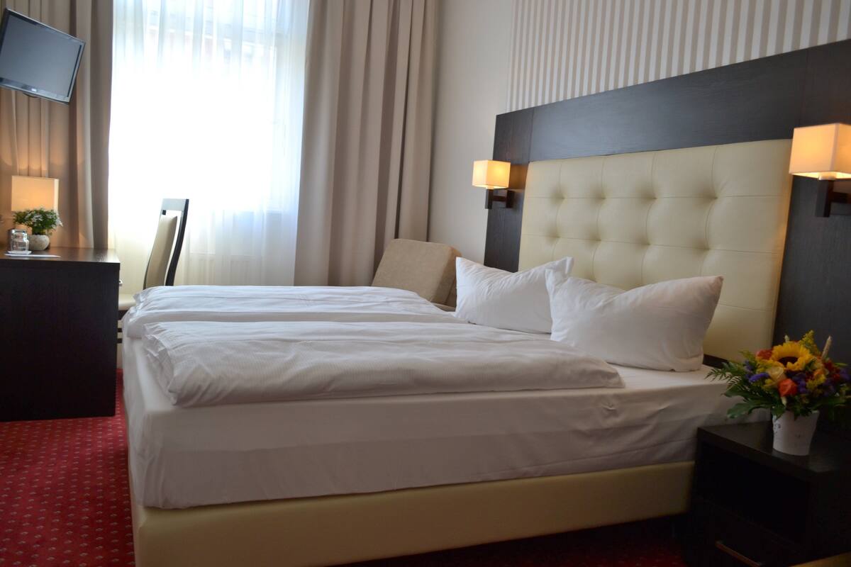 A hotel room with a large bed, a flat-screen TV, a desk, and a nightstand with a lamp and flowers.