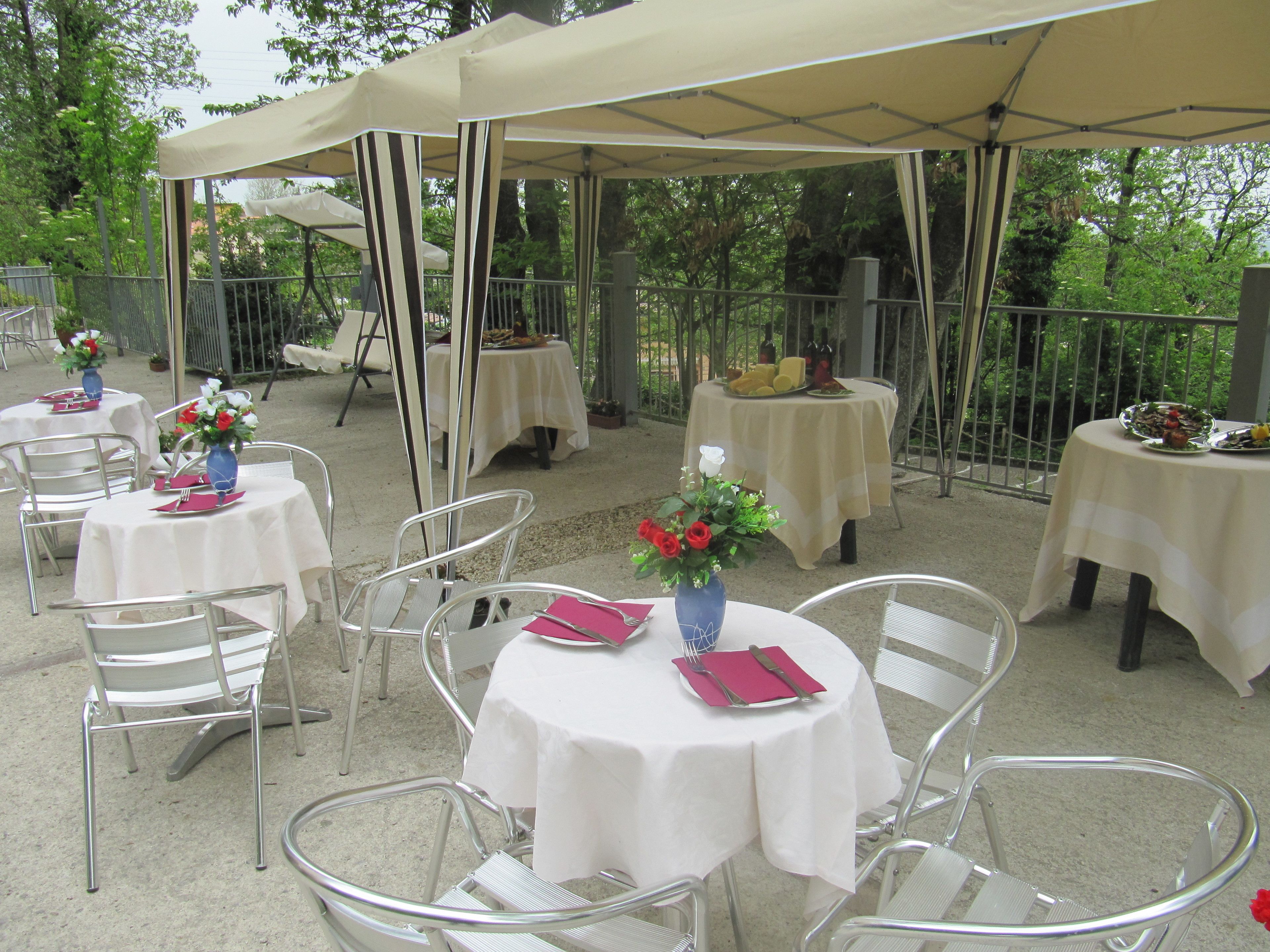 Un área de comedor al aire libre con mesas redondas cubiertas con manteles blancos, sillas de metal y sombrillas que brindan sombra.
