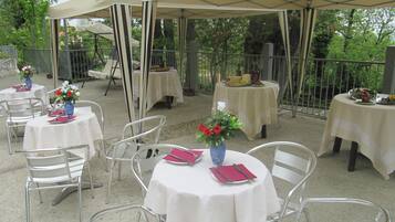 Un área de comedor al aire libre con mesas redondas cubiertas con manteles blancos, sillas de metal y sombrillas que brindan sombra.
