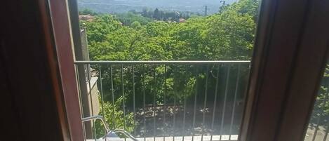A balcony with a metal railing, a chair, and a view of a green landscape with trees and distant mountains.