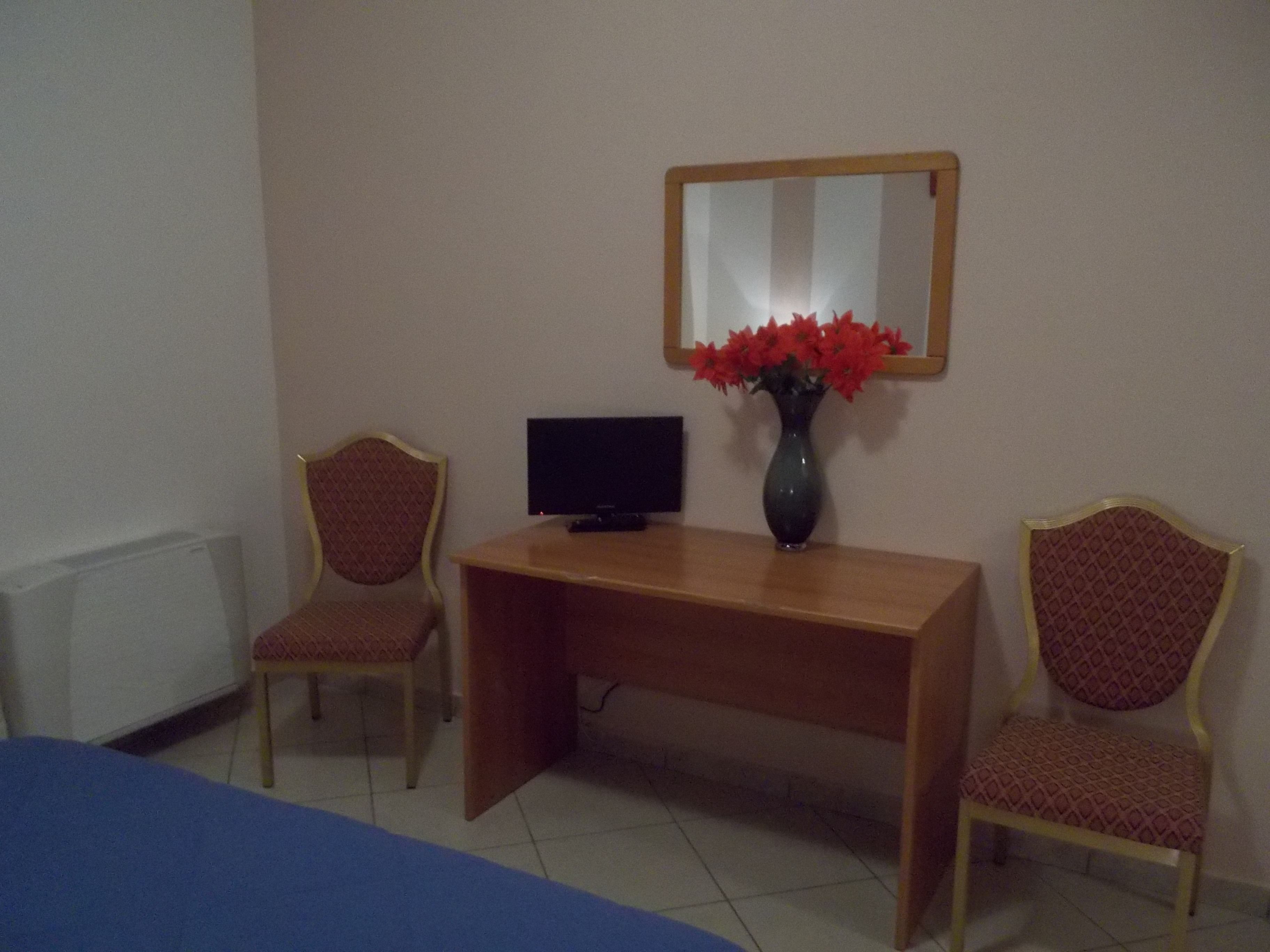 A room with a wooden desk, two patterned chairs, a vase with red flowers, a television, and a bed with a blue cover.