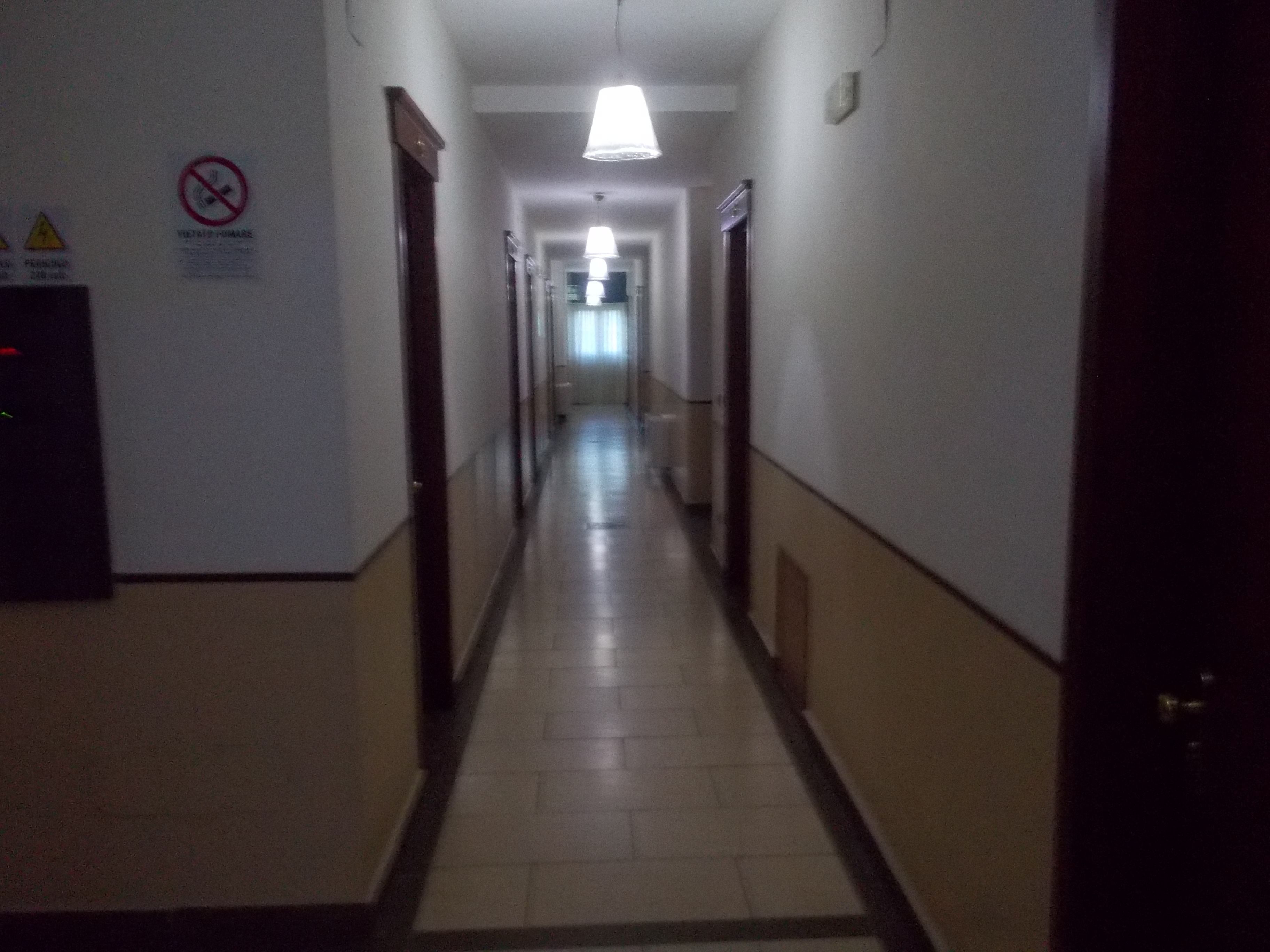 Un couloir étroit et faiblement éclairé, avec un sol carrelé et des portes de chaque côté.