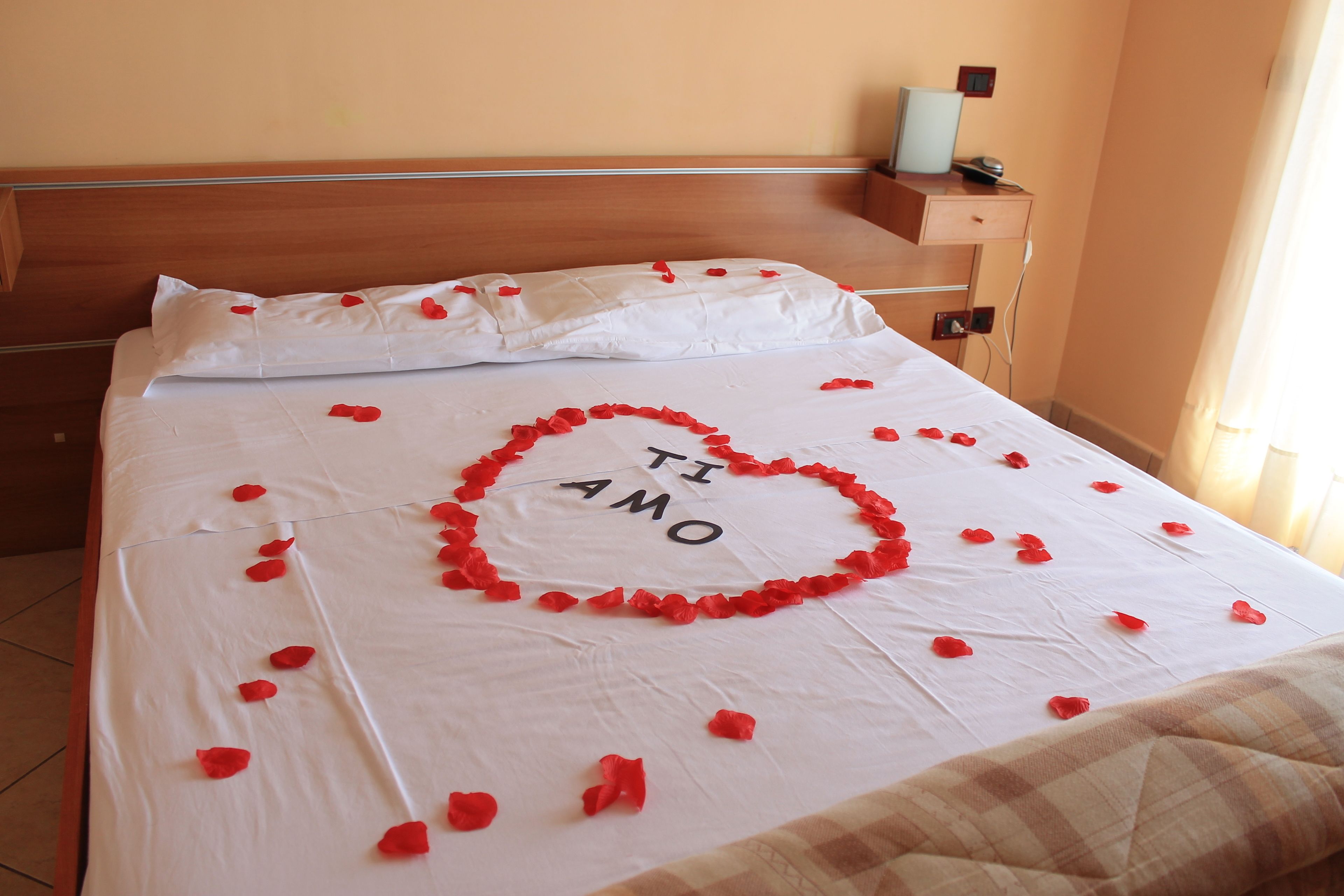 A bed with a heart-shaped arrangement of rose petals and the words "TI AMO" on the bedspread.