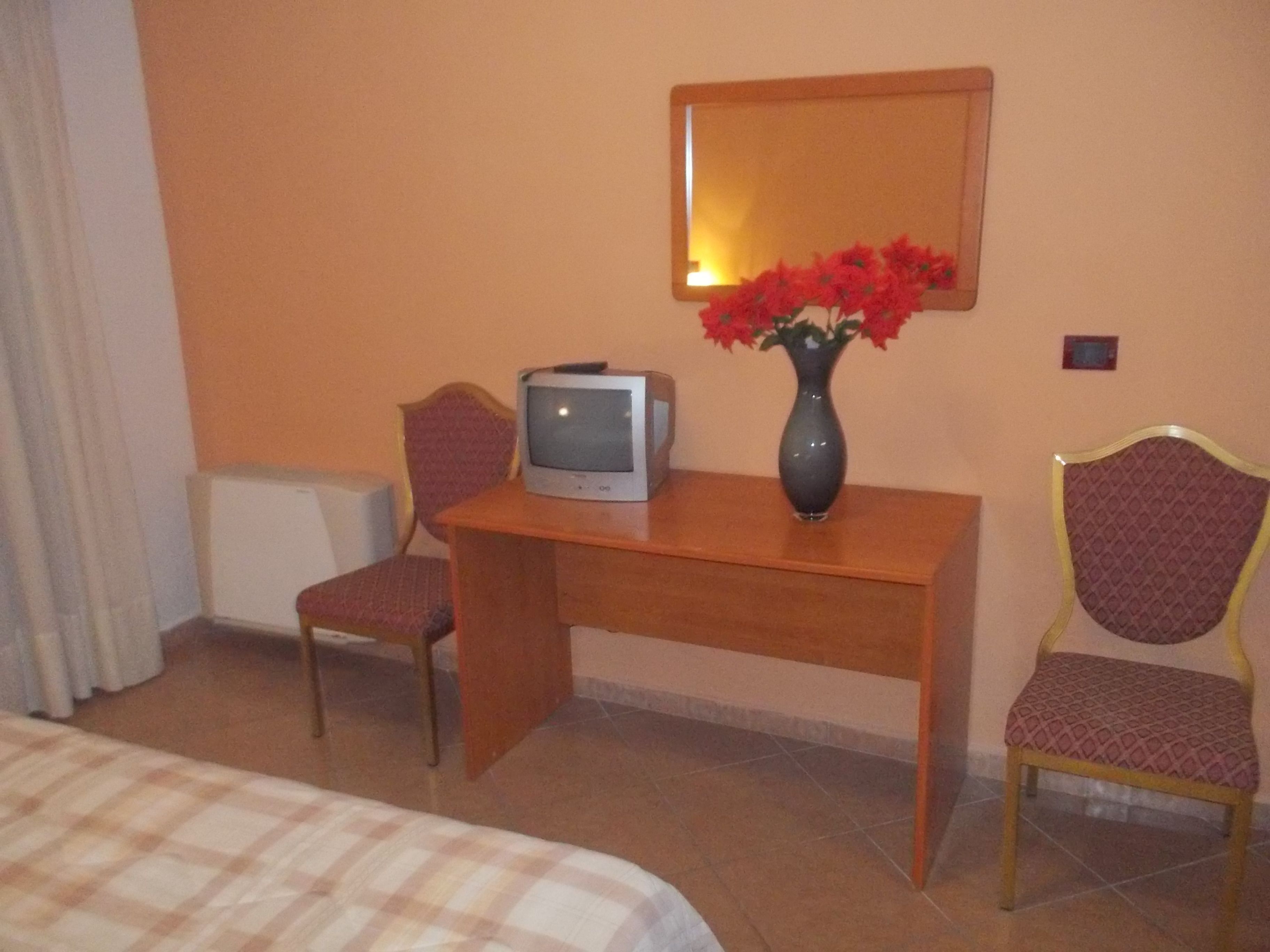 A hotel room with a bed, a desk with a television, a vase with flowers, and two chairs.