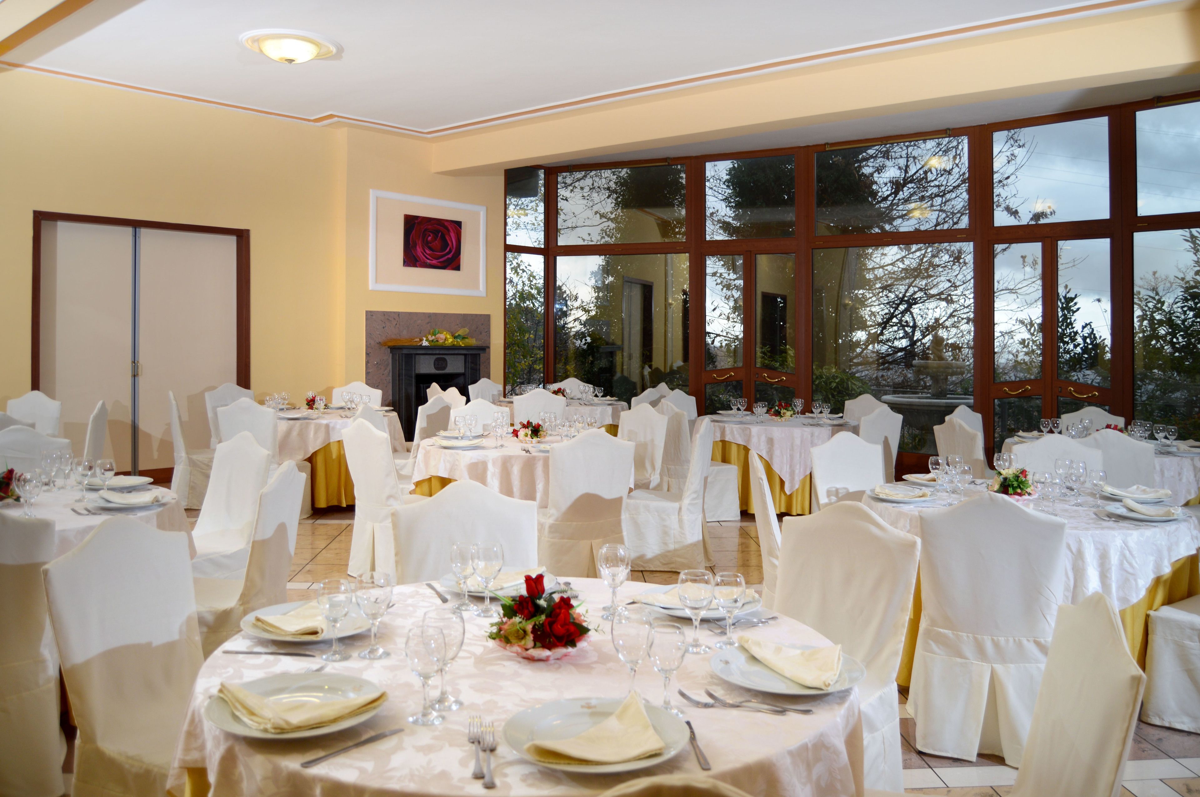 Una sala da banchetti con diversi tavoli rotondi apparecchiati per la cena, sedie bianche e ampie finestre che si affacciano su alberi e cielo.