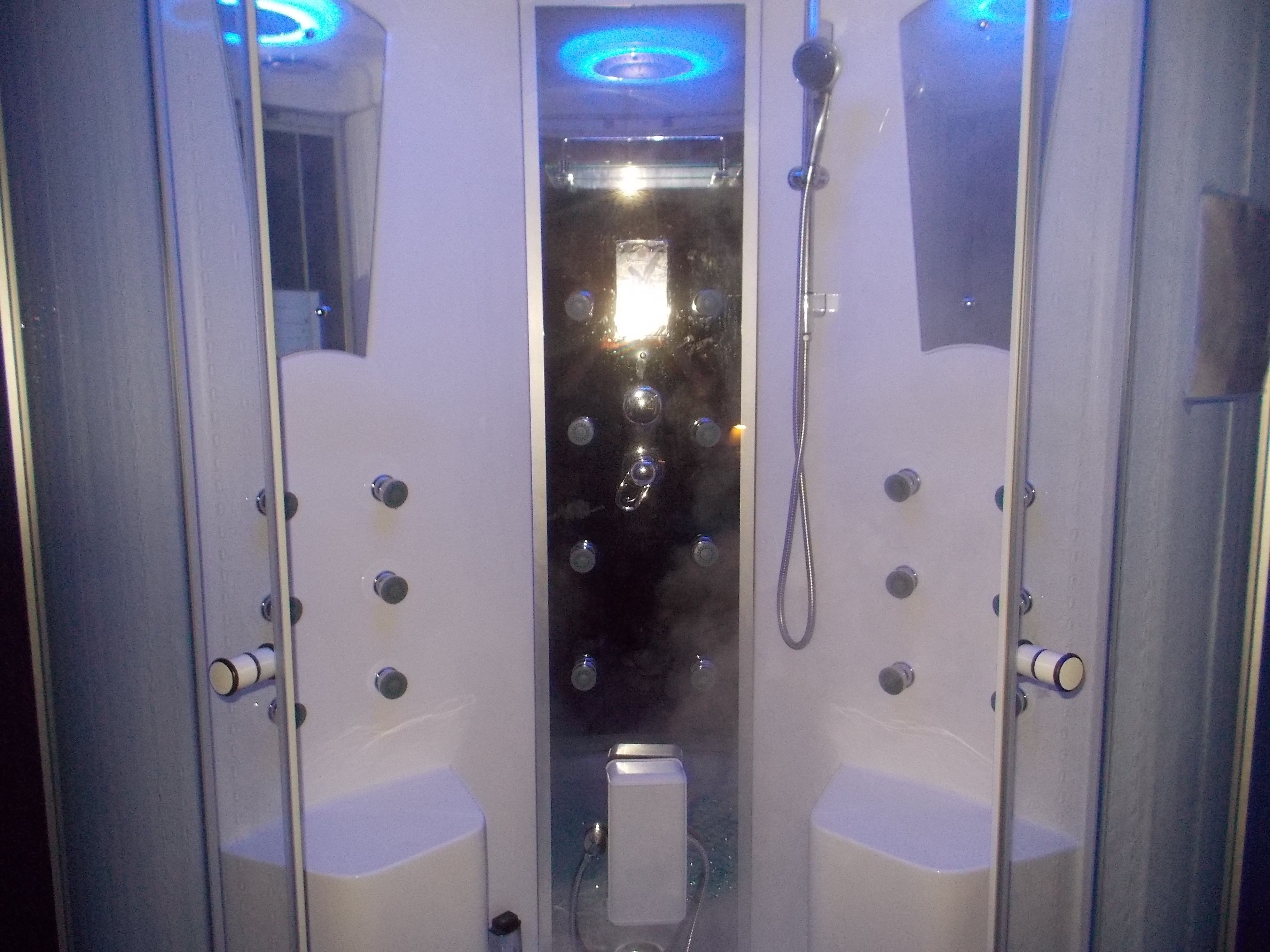 Cabina de ducha con paredes de vidrio, un cabezal de ducha manual, un panel de control con varios botones y un espejo en la puerta.