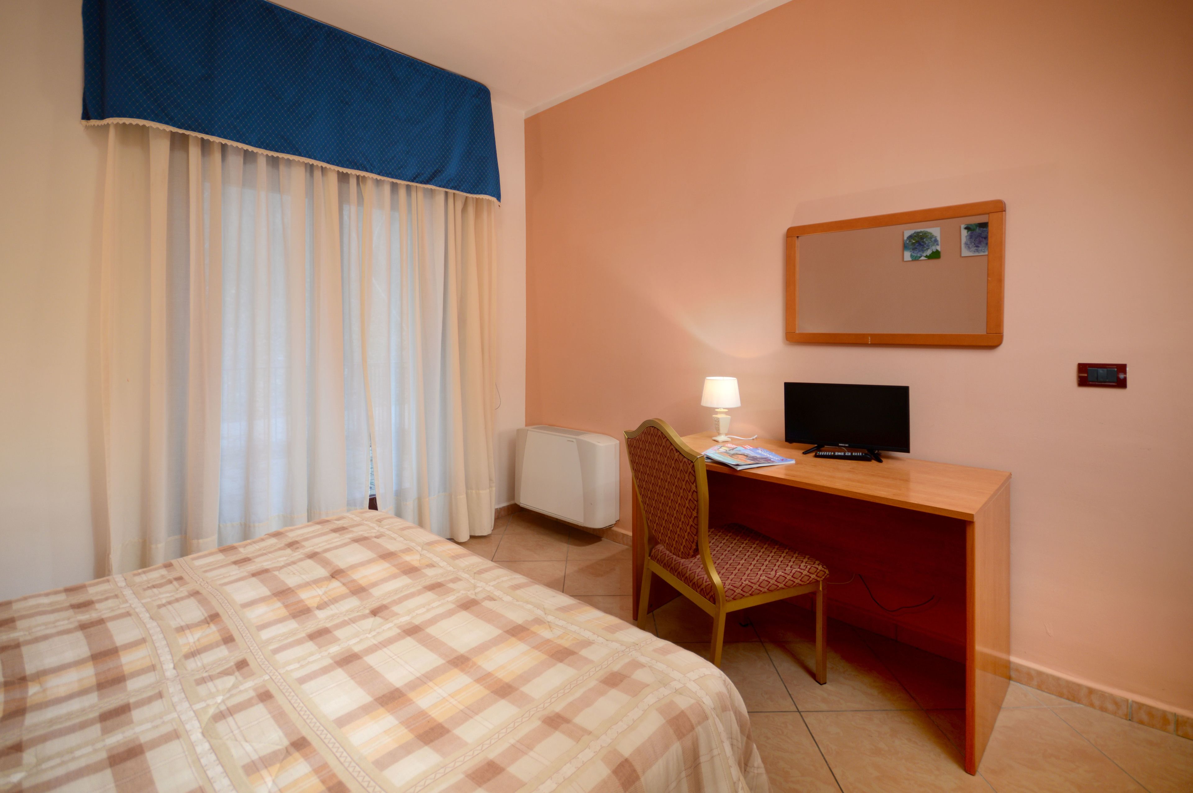 A hotel room with a bed, a desk with a computer, a chair, a lamp, and a wall-mounted mirror.