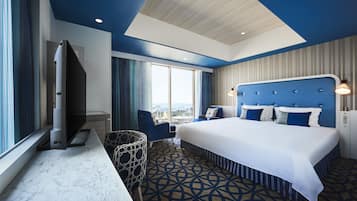 A hotel room with a large bed, a TV, a chair, and a view of the city.