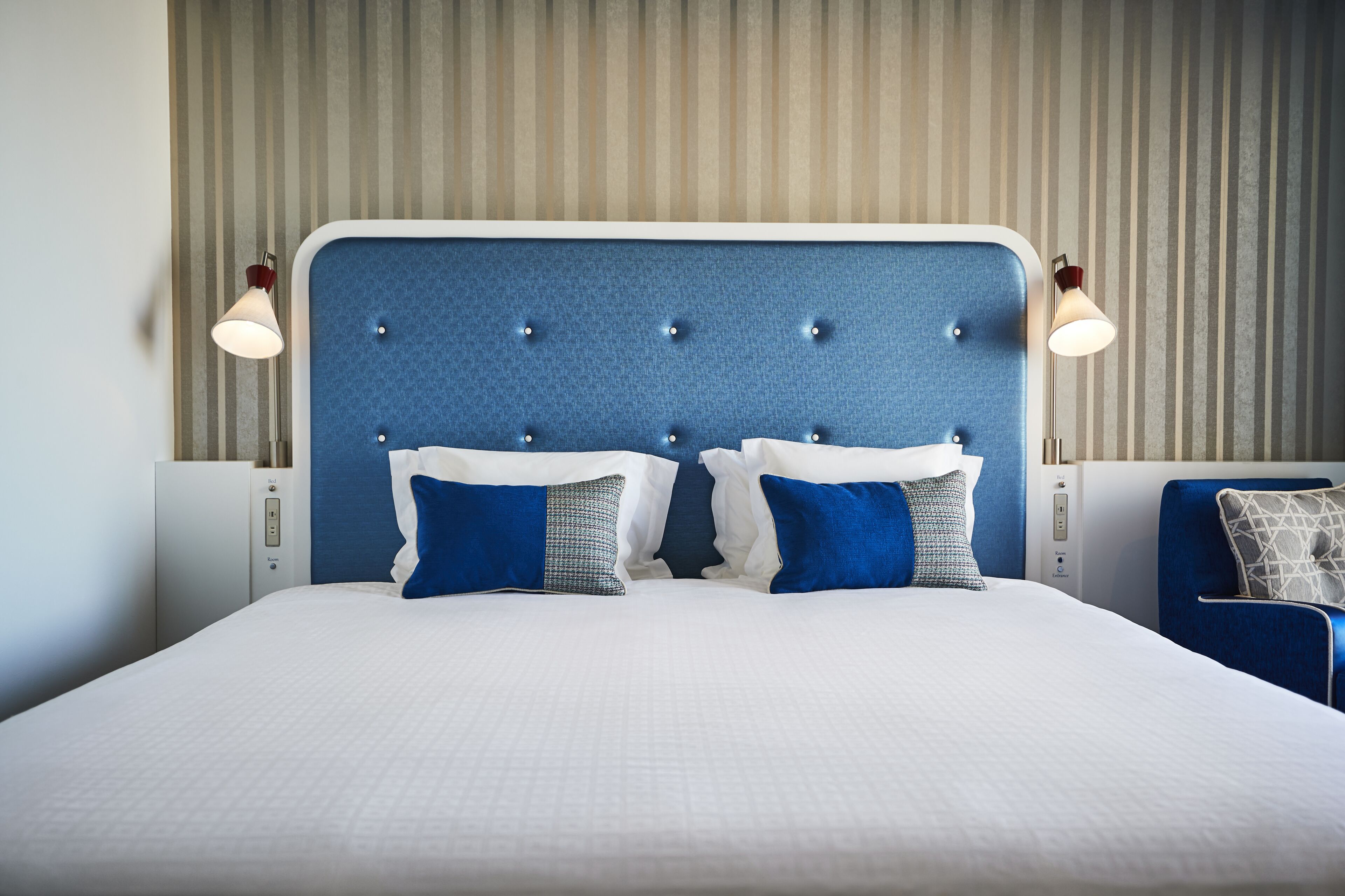 A neatly made bed with a blue headboard, two bedside lamps, and a blue chair.