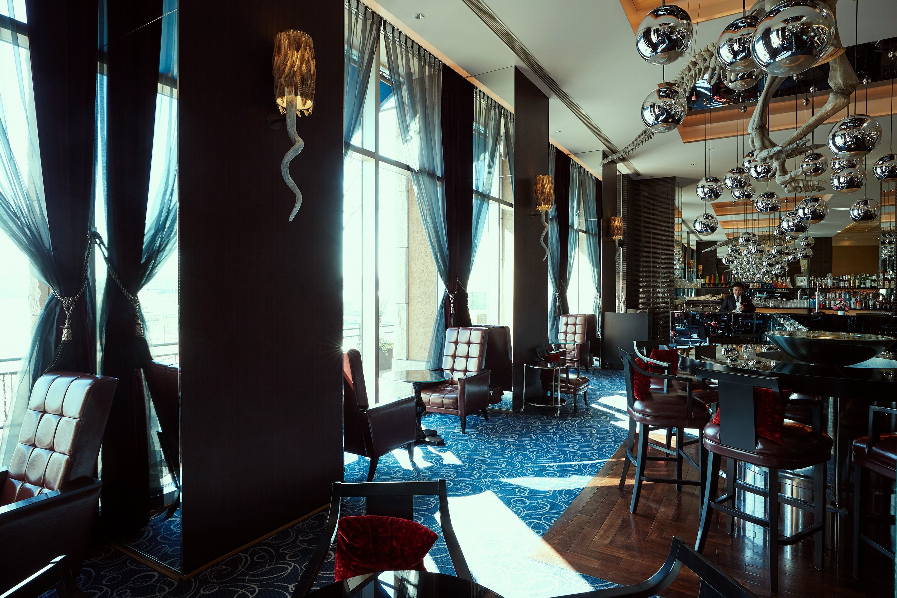 Un área de bar espaciosa con techos altos, ventanas amplias y una decoración moderna que incluye espejos colgantes y una alfombra estampada.