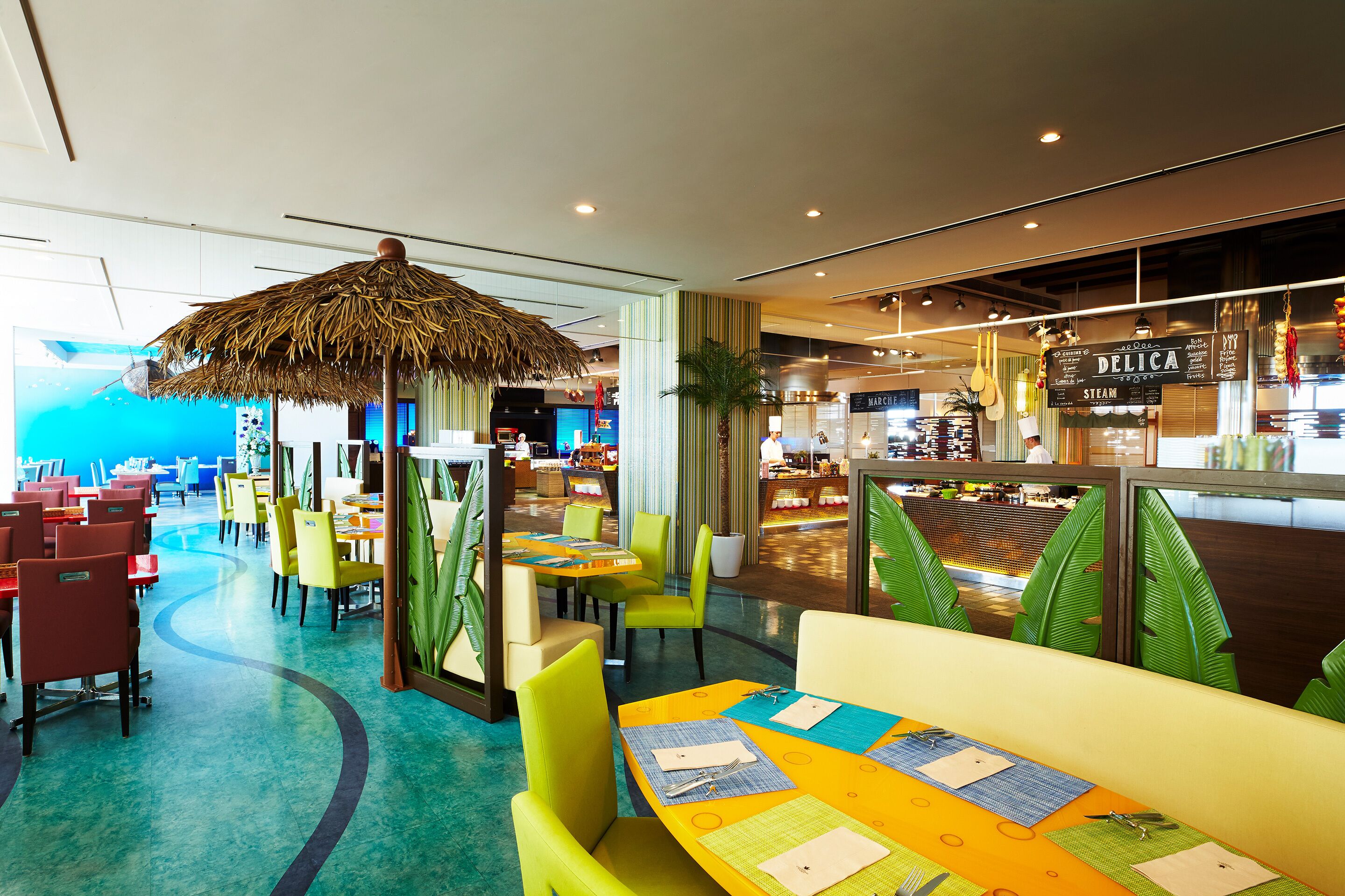 Un restaurant avec des chaises et des tables aux couleurs vives, un parasol en chaume et une vue sur l’espace cuisine.