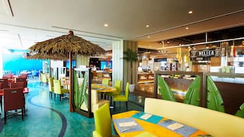 Un restaurant avec des chaises et des tables aux couleurs vives, un parasol en chaume et une vue sur l’espace cuisine.