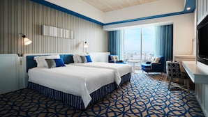 Ein Hotelzimmer mit zwei Betten, einem Schreibtisch, einem Stuhl, einem Fernseher und einem großen Fenster mit Blick auf die Stadt.