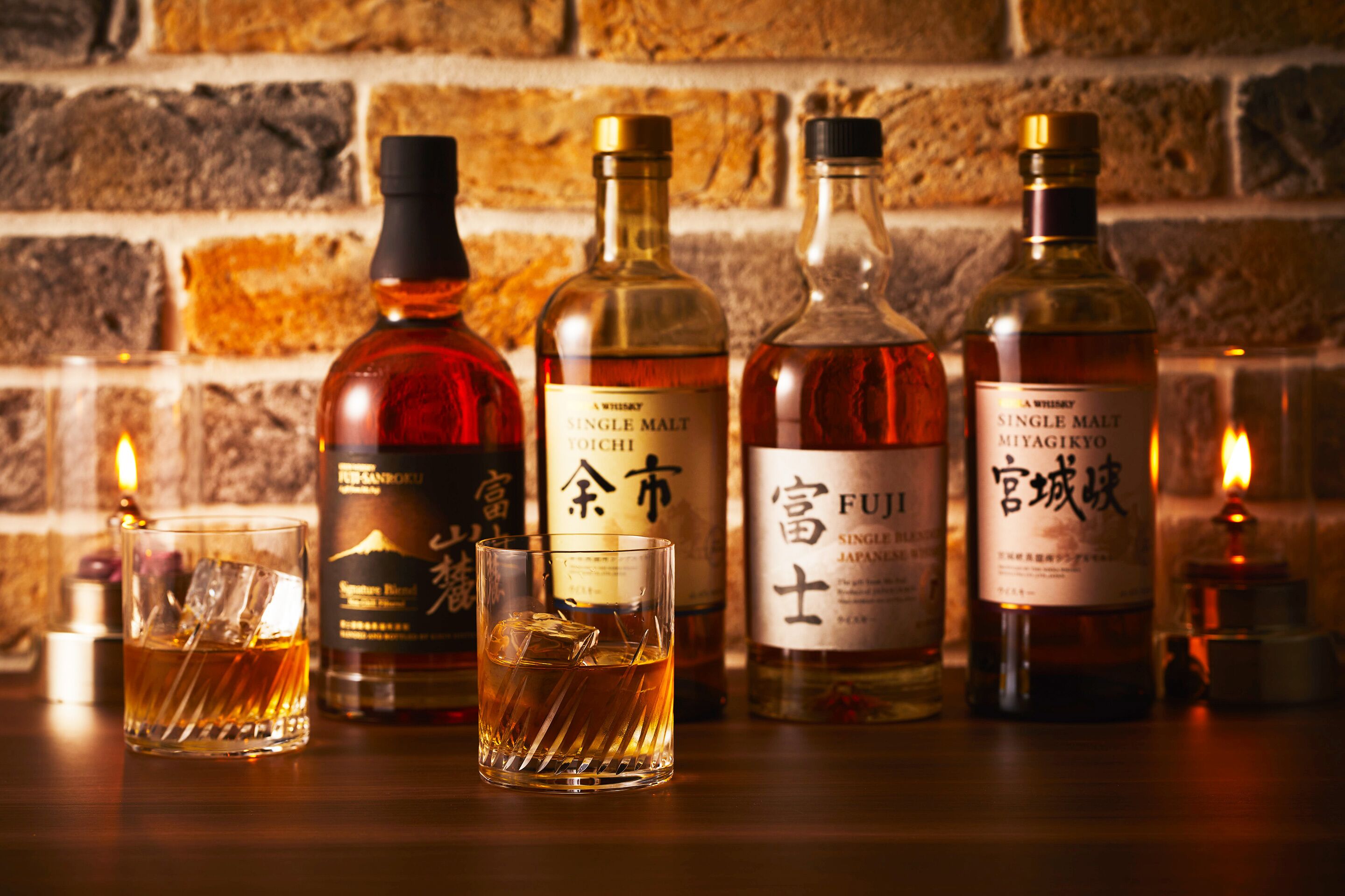 A selection of whiskey bottles and glasses on a wooden surface with a brick wall background.