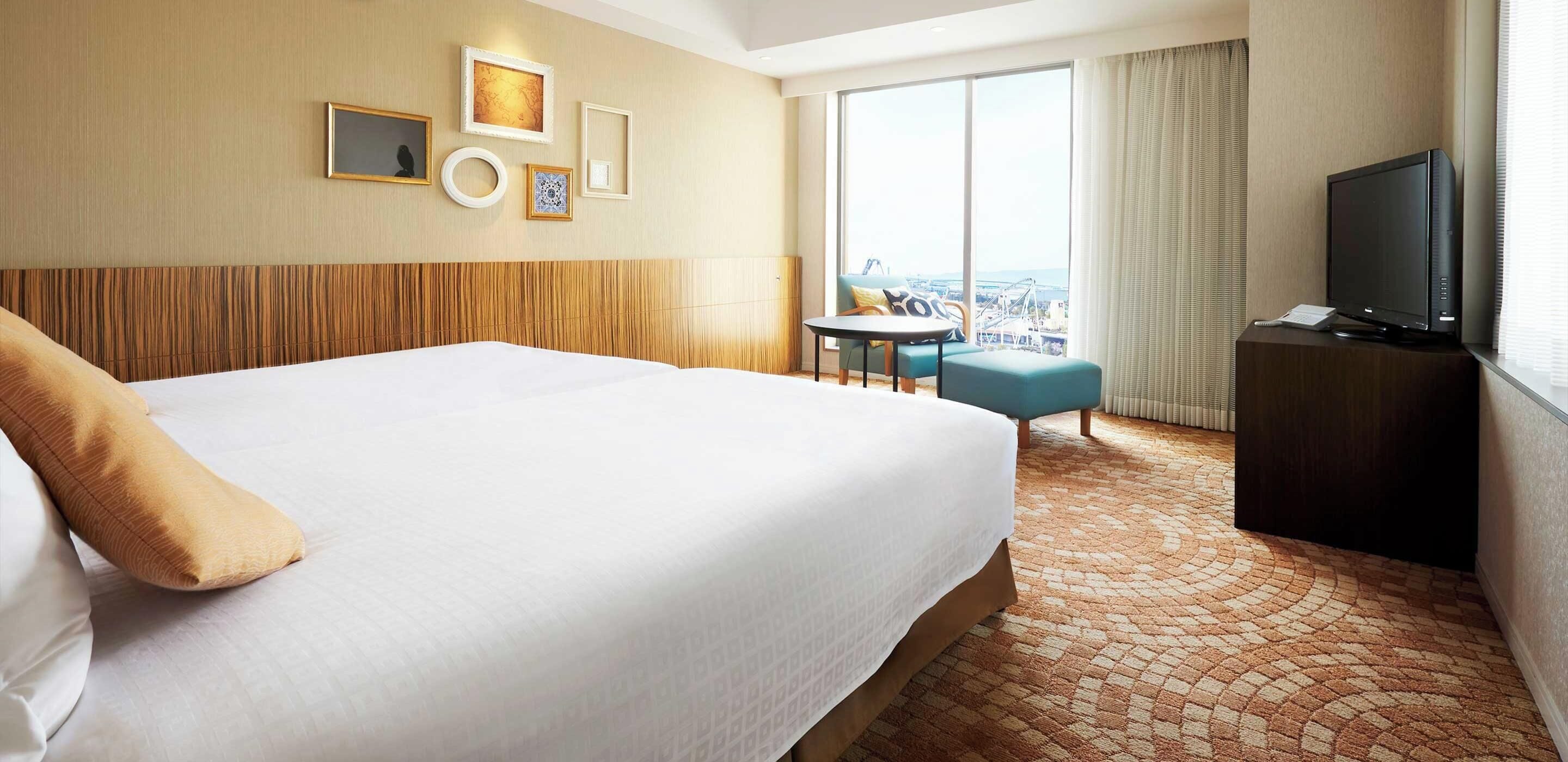 A hotel room with a large bed, a television, a small table, and a view of a cityscape through the window.