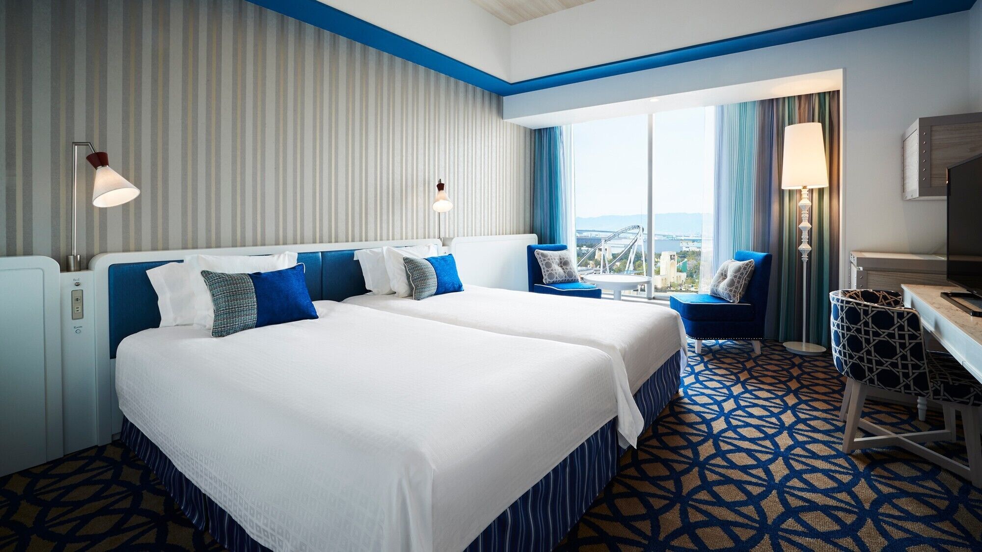 Habitación de hotel con dos camas, un escritorio, una silla y un amplio ventanal con vistas.